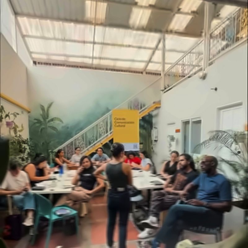 A cultural gathering in the courtyard: people seated around tables listen to someone standing under the translucent roof.