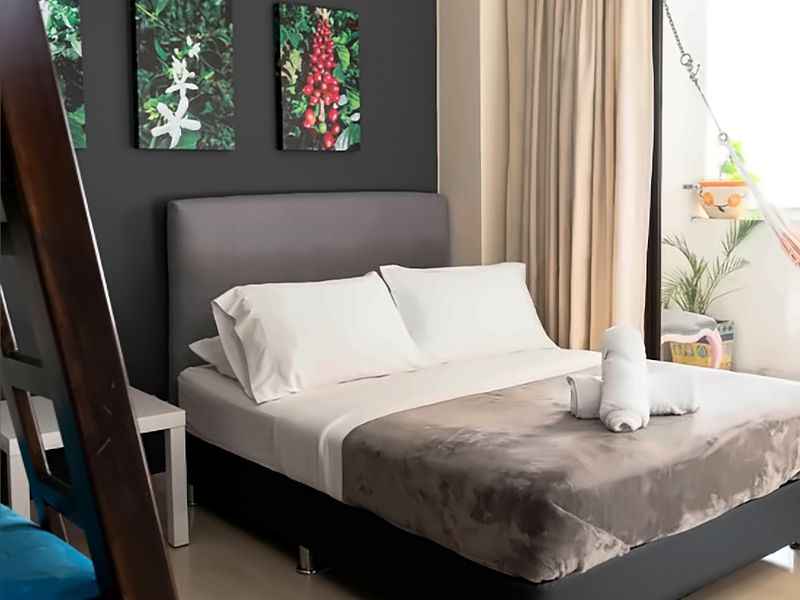 Habitación Castillo: cama doble con cubrelecho gris, pared oscura con cuadros de flor y fruto de café, litera de madera a la izquierda y balcón con hamaca.