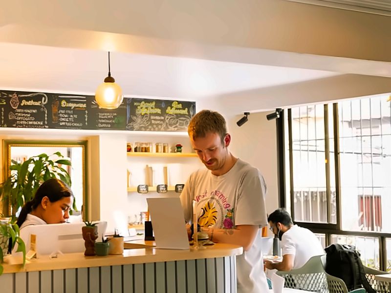Barra del café con tablero de menú y lámpara colgante; personas trabajan con portátiles junto al ventanal.