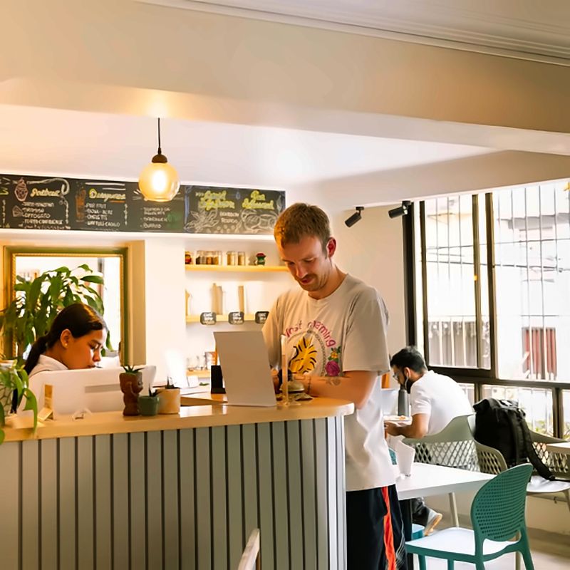 Barra del café con tablero de menú y lámpara colgante; personas trabajan con portátiles junto al ventanal.