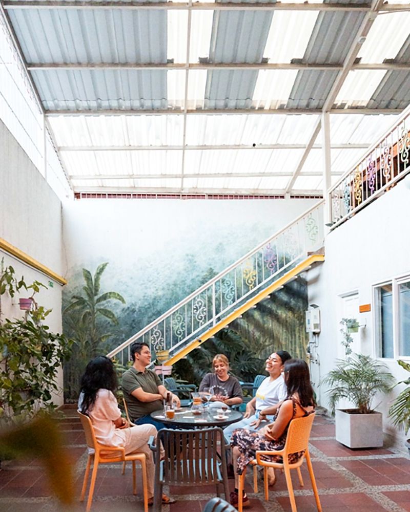 Patio cubierto de Casa Azul: un grupo conversa alrededor de una mesa bajo el mural de vegetación y la escalera blanca de baranda ornamental con borde amarillo.