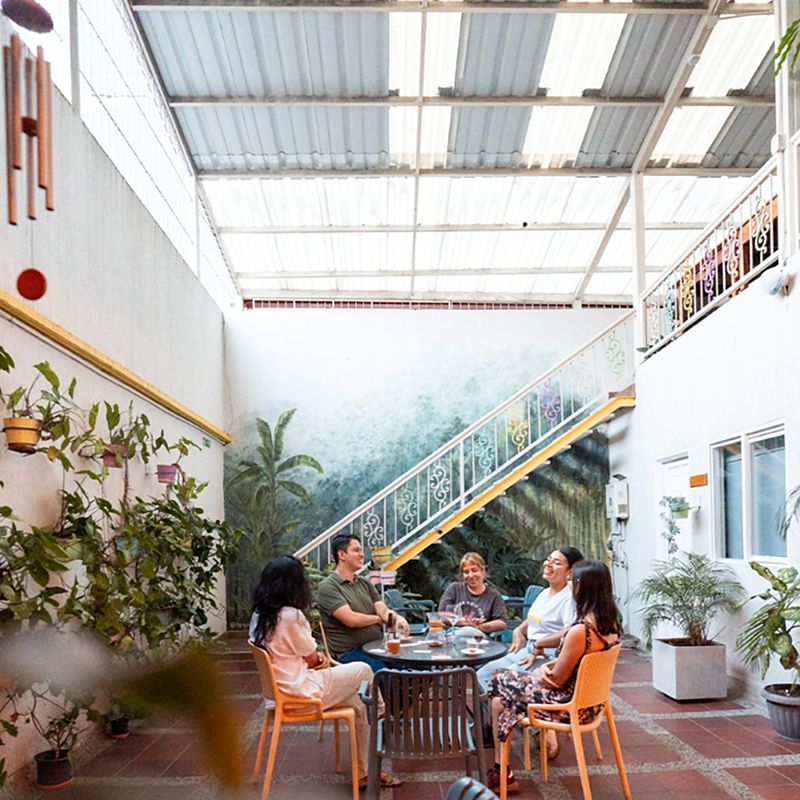 Patio cubierto de Casa Azul: un grupo conversa alrededor de una mesa bajo el mural de vegetación y la escalera blanca de baranda ornamental con borde amarillo.