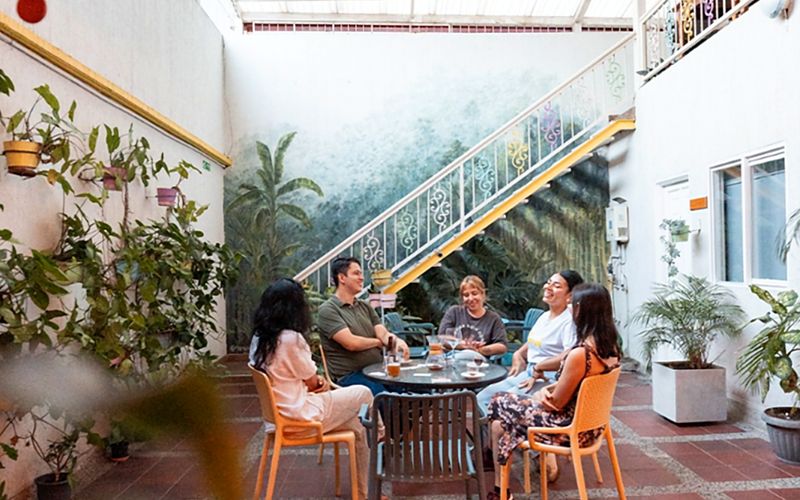 Patio cubierto de Casa Azul: un grupo conversa alrededor de una mesa bajo el mural de vegetación y la escalera blanca de baranda ornamental con borde amarillo.
