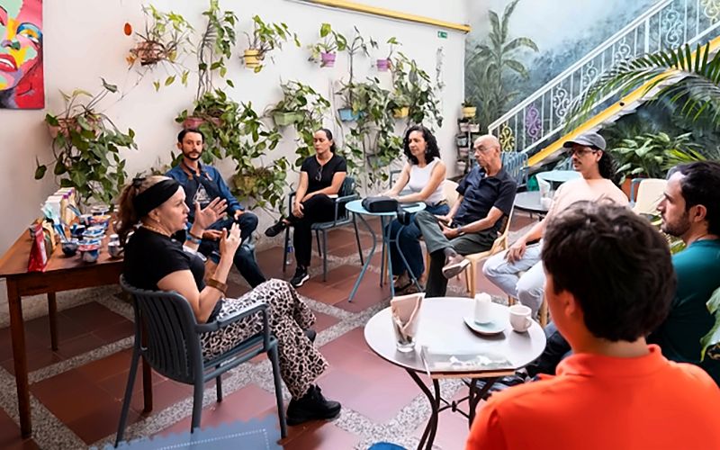 Encuentro en el patio: un grupo grande conversa sentado en círculo entre matas colgantes y el mural.