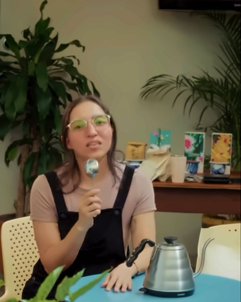 Charla de café: una mujer habla con una cuchara de catación en la mano, junto a una tetera de cuello de cisne sobre una mesa turquesa.