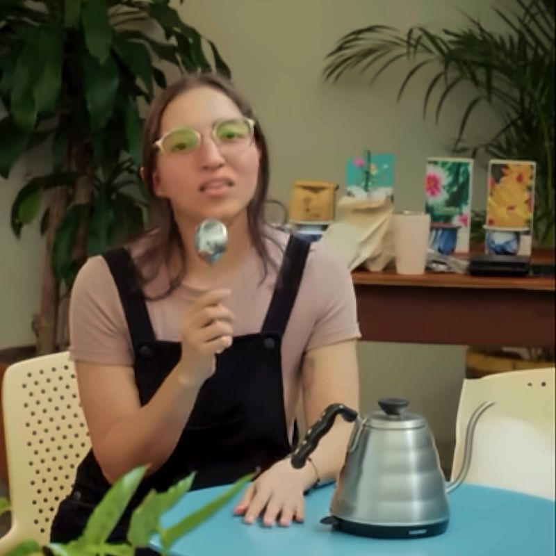 Charla de café: una mujer habla con una cuchara de catación en la mano, junto a una tetera de cuello de cisne sobre una mesa turquesa.