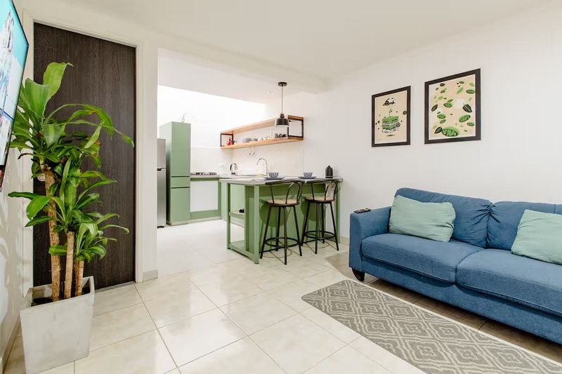 Apto: sala con sofá azul, cocina con gabinetes verdes y barra con taburetes.