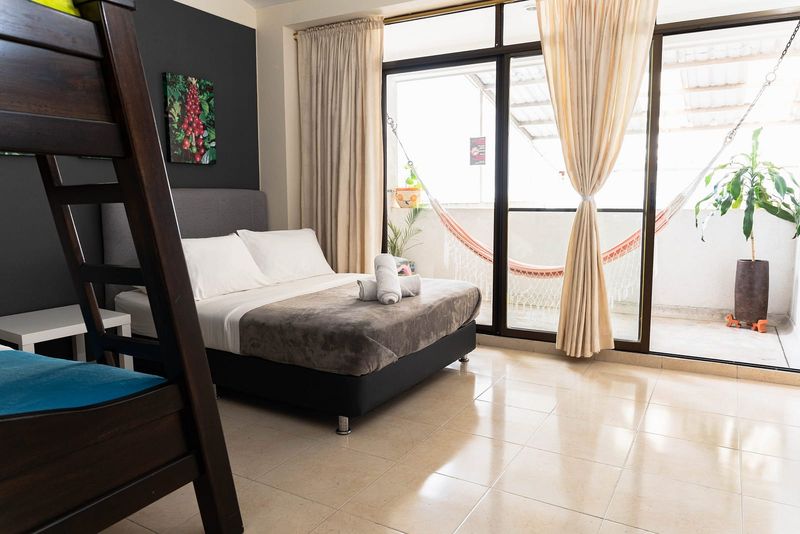 Habitación Castillo: litera de madera, cama doble con cubrelecho gris, pared oscura con cuadro de café y puerta al balcón con hamaca.