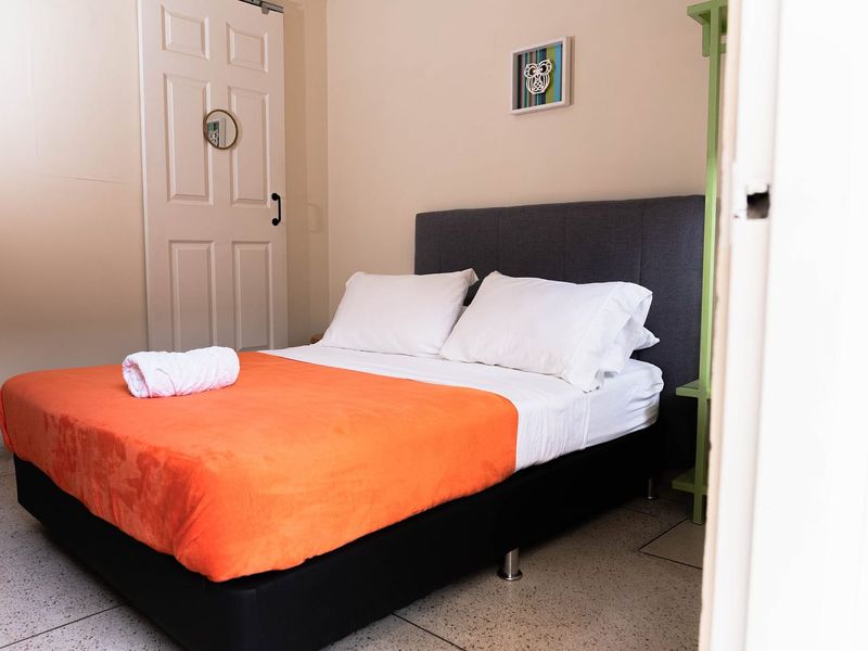 Habitación Tabi: cama doble con cobija naranja, cabecero gris y puerta blanca.
