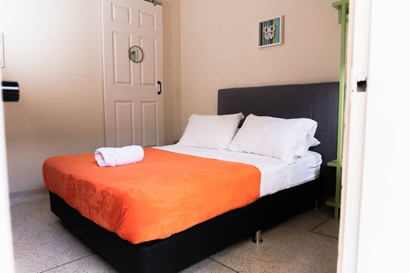 Habitación Tabi: cama doble con cobija naranja, cabecero gris y puerta blanca.